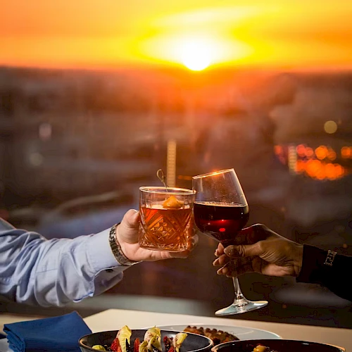 Two people toast with cocktails over a sunset dinner&mdash;glasses raised above bowls of food as the warm sky glows in orange hues.