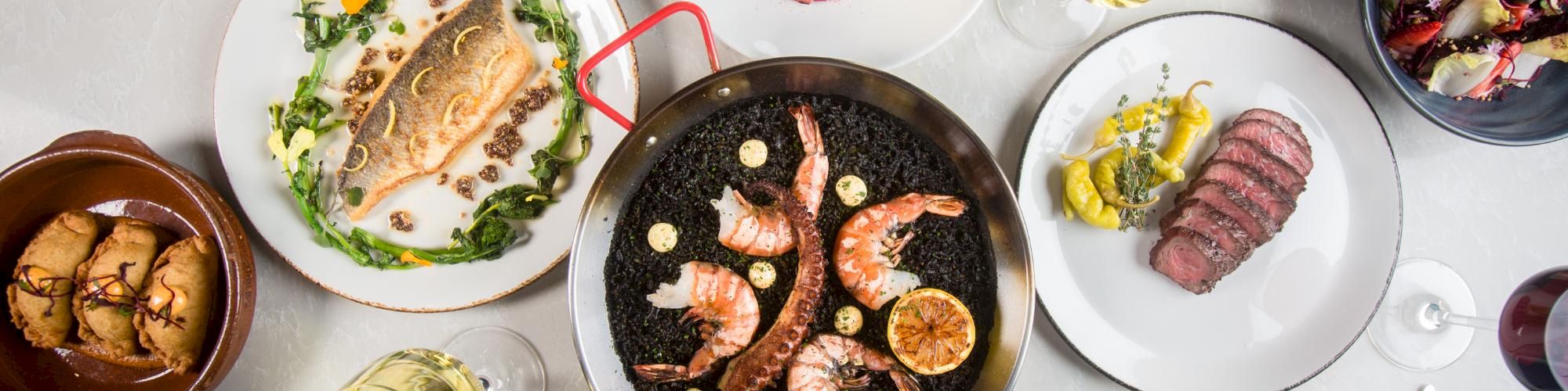 A top-down view of a table loaded with seafood and meats: shrimp on a grill pan, breaded items, sliced steak, pasta, and assorted sides, with wine glasses.