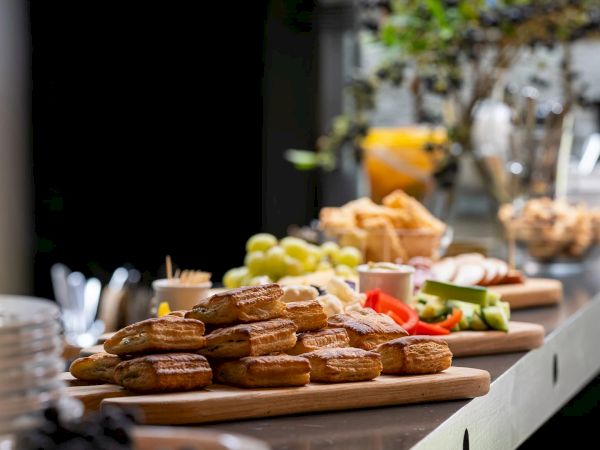 A row of pastries and fruit spread along a buffet counter, with croissants, &eacute;clairs, grapes, and assorted sliced fruit on wooden boards.