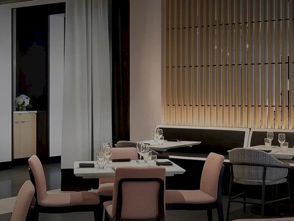 A chic restaurant interior with round and square tables, pink upholstered chairs, subtle lighting, and a decorative wall pattern on the left.
