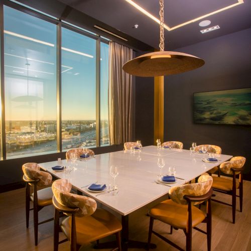 A stylish dining room with a long wooden table, eight chairs, warm lighting, and large windows showing a sunset view.