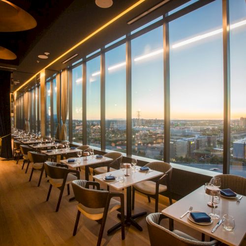 A cozy, modern cafe with warm wood accents, large windows, and comfy seating along a long bar overlooking a cityscape at sunset.