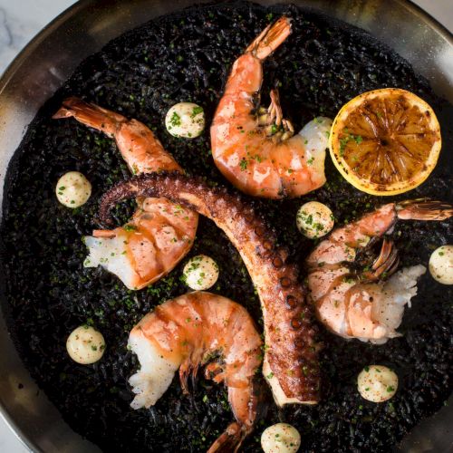 A round, black-framed dish with a skillet of fried eggs, sausages, and hash browns, garnished with a few herbs and a sunny yellow yolk.