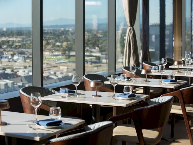 A stylish, sunlit restaurant with white tables, modern chairs, wine glasses, and a panoramic city view through large windows, ready for diners.