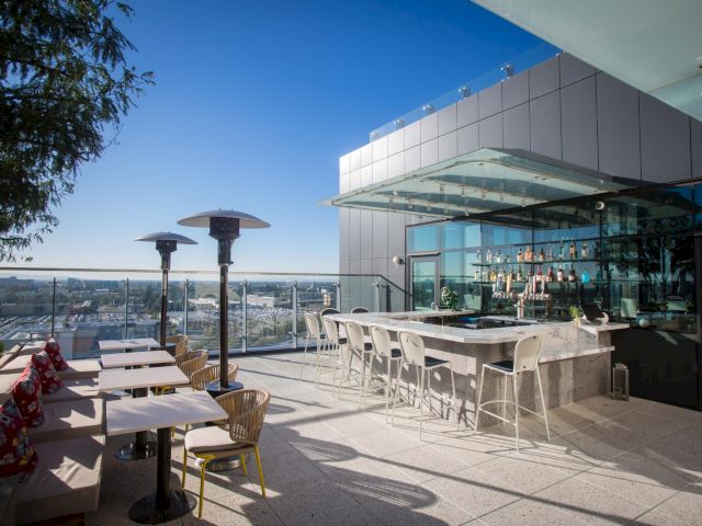 A chic rooftop bar with outdoor seating, a long bar counter, and city views under a clear blue sky.