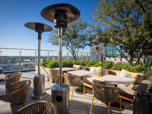 Rooftop patio with wicker chairs, beige couches, and yellow cushions, potted plants, and tall patio heaters under a clear blue sky.
