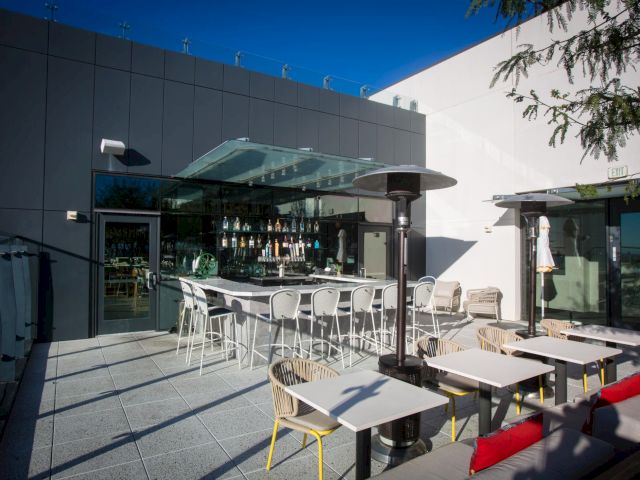 An outdoor bar and seating area with white tables and chairs, a shaded bar counter, and a heater, in a modern patio setting.