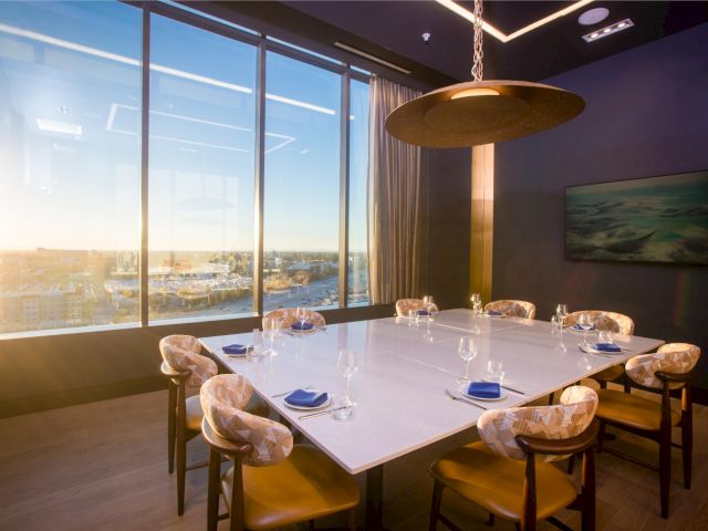 A modern dining room with a long white table set for twelve, glassware, and napkins, by a wall of floor-to-ceiling windows at sunset.