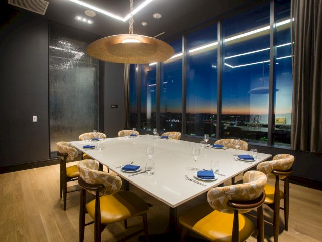 A modern dining room with a glass-top square table set for eight, gold chairs, a large pendant light, and floor-to-ceiling windows at dusk.