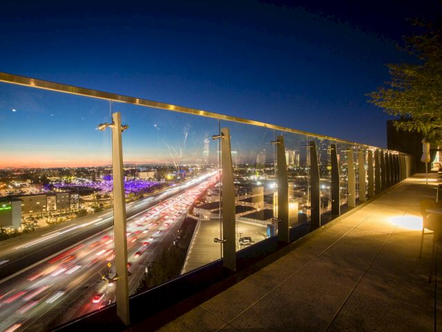 City skyline at dusk with blurred car lights on a highway below, viewed from a glass railing walkway; warm lights along the path glow softly.