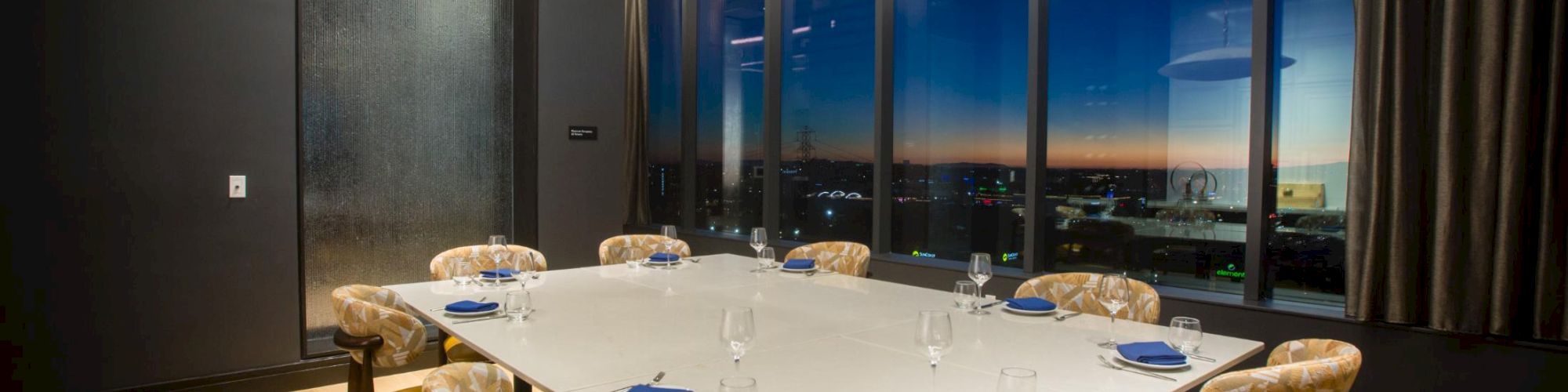 A modern private dining room with a large white square table, yellow cushioned chairs, blue napkins, glass walls, and a city view at dusk, stylish lighting.