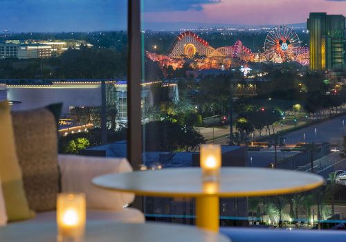 A cozy indoor lounge with candles on a round table, glass windows, and a nighttime cityscape with a lit Ferris wheel outside. Top it at 140 characters, always ending the sentence.